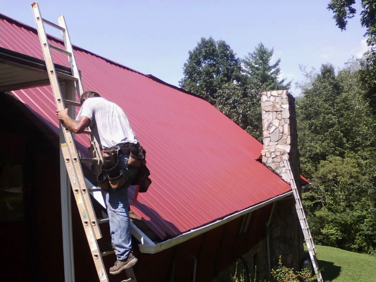 Roofer on a ladder beside a red metal roof and stone chimney — Soffit & Fascia Repair by Roofing Brothers in Wrightstown